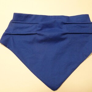 May include: A blue bandana-style bib with a white, fuzzy lining. The bib has a folded top with two horizontal seams. The bib is made of a soft fabric and is designed to be worn around the neck.
