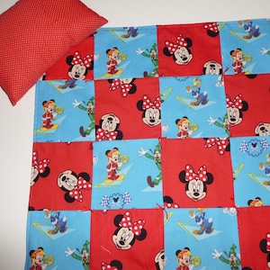 May include: A red polka dot pillow rests beside a patchwork quilt. The quilt features a pattern of red and blue squares with images of Minnie Mouse, Donald Duck, and Goofy. The quilt is a children's item.