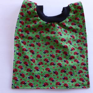 May include: A green bib featuring a repeating pattern of red ladybugs and small red hearts. The bib has a solid black fabric neckline and a rectangular shape with rounded bottom corners, constructed with visible stitching along the edges.
