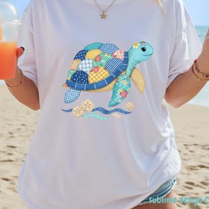 Puede incluir: Camiseta blanca con un diseño de tortuga patchwork colorido. La tortuga tiene una cabeza azul y aletas amarillas, con un caparazón hecho de varias telas estampadas. El diseño incluye olas y conchas marinas. El texto "sublime design CStudio" está en la parte inferior.