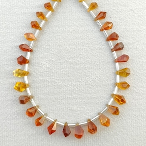 Puede incluir: Un collar de piedras preciosas facetadas en forma de lágrima, en tonos amarillos, naranjas y marrones. Las cuentas están ensartadas en un alambre plateado, creando un efecto degradado. Las piedras están espaciadas uniformemente.