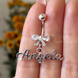 May include: A silver belly button ring with a bow-shaped charm and the name "Angela" in a cursive font. The ring has a small ball at the top and is held up by a hand.