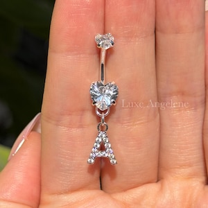 May include: A silver belly button ring with a clear, heart-shaped gem in the center and a sparkling letter "A" pendant. The top gem is a clear, round shape. The jewelry is held up by a person's hand.
