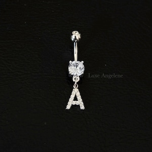 Puede incluir: Un piercing para el ombligo plateado con una gema redonda y transparente grande y una letra "A" colgante incrustada con piedras transparentes más pequeñas. El texto "Luxe Angelene" es visible en el fondo.