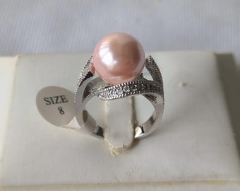 May include: A silver ring featuring a large, round, pink pearl. The ring band has a decorative design with small, clear stones. A size 8 tag is attached to the ring.