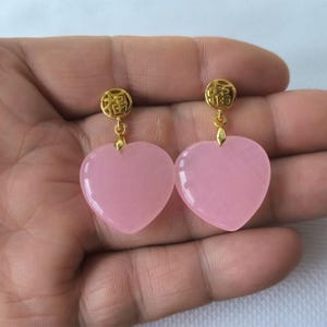 Peut inclure: Une paire de boucles d'oreilles pendantes composées de pierres lisses en forme de cœur rose clair, suspendues à des clous circulaires dorés. Chaque clou est gravé d'un caractère chinois distinct, et les cœurs pendent librement du métal.