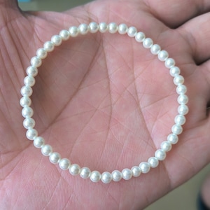 Puede incluir: Un brazalete de perlas blancas con un diseño sencillo. El brazalete está hecho de pequeñas perlas redondas que están enhebradas en una cuerda fina.