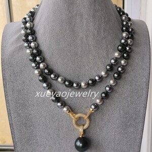 May include: A double-strand necklace featuring alternating dark gray and light gray spherical beads. The necklace includes a gold-toned circular clasp encrusted with small clear stones, from which a single large dark gray bead hangs. Text reads xueyaojewelry.