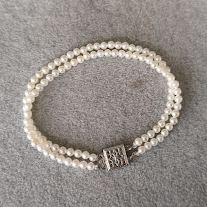 Può includere: Un braccialetto di perle a due fili con una chiusura quadrata color argento. Il braccialetto presenta piccole perle bianche rotonde. La chiusura ha un design traforato decorativo. Il braccialetto è esposto su una superficie grigia.