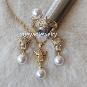 Puede incluir: Un conjunto de joyas de tono dorado que incluye un collar, pendientes y un anillo. Cada pieza tiene un pequeño diseño de cabeza de pantera detallado con ojos verdes y está adornado con diamantes de imitación transparentes. El collar y los pendientes también presentan una gota de perla blanca.