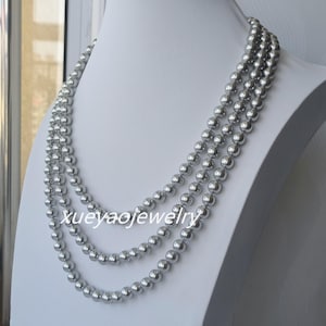 Puede incluir: Tres hileras de collares de perlas grises sobre un maniquí blanco. Las perlas son redondas y de tamaño uniforme, creando un aspecto clásico y elegante. El texto "xueyaojewelry" es visible.