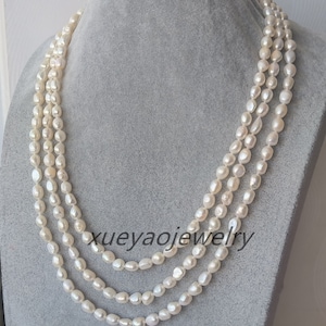 Op de afbeelding: Een drieregenige parelketting met witte, ovale parels. De ketting wordt getoond op een grijze mannequin.