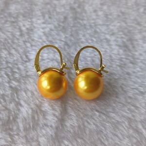 May include: A pair of gold-colored hoop earrings with large, round, golden-yellow pearls. The earrings have a simple, elegant design, with a hinged closure. The pearls have a smooth, lustrous surface, and the gold metal has a polished finish.