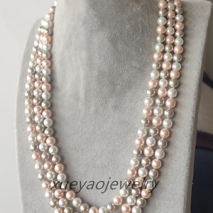 Könnte beinhalten: Eine mehrreihige Perlenkette mit drei Reihen Perlen in den Farben Weiß, Creme und Blush-Rosa. Die Kette ist aus Kunstperlen gefertigt und etwa 46 cm lang.