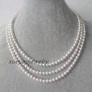 Puede incluir: Un collar de perlas de tres hileras con perlas blancas. El collar está sobre un fondo gris.