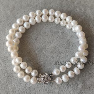 Puede incluir: Un brazalete de perlas blancas con una doble hilera de perlas y un cierre plateado. El brazalete está sobre una superficie gris.