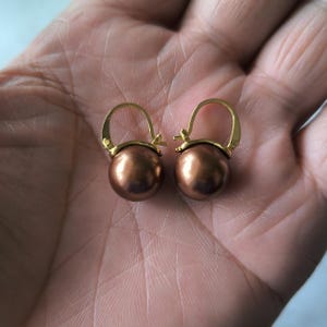 Pode incluir: Um par de brincos esféricos cor de cobre com argolas douradas. Os brincos são redondos e têm um brilho metálico. As argolas têm um design simples e curvo.