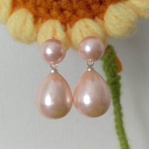 Può includere: Un paio di orecchini pendenti caratterizzati da una perla rotonda e liscia di colore rosa chiaro come perno, collegata tramite un piccolo anello in metallo argentato a un pendente a goccia più grande, anch'esso di colore rosa chiaro.