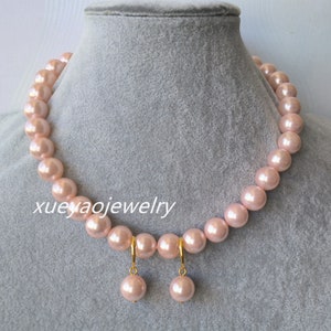 Puede incluir: Un collar de perlas rosa rubor y un juego de pendientes a juego. El collar presenta grandes perlas redondas ensartadas. Los pendientes tienen un aro dorado y una sola perla a juego. El texto "xueyaojewelry" es visible.