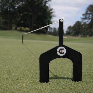 Puede incluir: Una herramienta de alineación de golf negra con una cuerda de alineación blanca y roja. La herramienta tiene una base curva con un logotipo y un poste vertical. La herramienta está en un campo de golf verde.