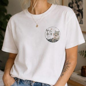 Op de afbeelding: Een wit T-shirt met korte mouwen en een ronde afbeelding op de linkerborst. De afbeelding toont een anime-figuur met kattenoren die op een reling leunt, met de Japanse tekst ヤニネコ en de Engelse tekst YANI NEKO.