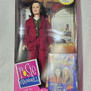 May include: A Rosie O'Donnell doll in a red blazer and matching trousers with a green pinstriped vest, packaged in a box with a Rosie O'Donnell Activity Zone booklet. The box features the text Rosie O'Donnell Friend of Barbie and a QVC sticker.