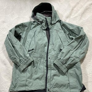 May include: A sage green Marmot hooded jacket with a black interior and trim. The jacket has a zipper closure, snap buttons, and elastic cuffs. The Marmot logo is visible on the chest.