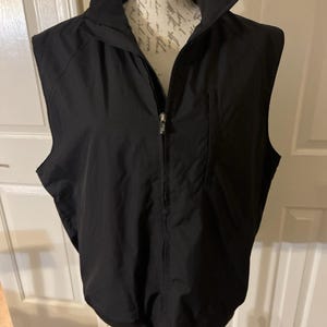 May include: Black vest with a full-length zipper and a stand-up collar. The vest has a streamlined design with a pocket on the left side. The vest is displayed on a mannequin.