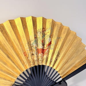 May include: A decorative folding fan with a gold leaf design and a black wooden handle. The fan features a painted illustration of a figure in traditional attire. The fan is displayed on a black stand.
