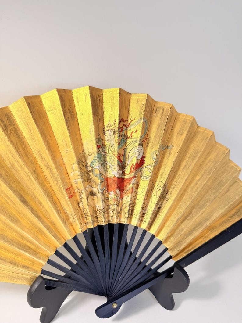 May include: A decorative folding fan with a gold leaf design and a black wooden handle. The fan features a painted illustration of a figure in traditional attire. The fan is displayed on a black stand.