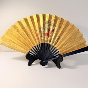 May include: A decorative folding fan with a gold-colored surface and a black handle. The fan features an ornate design with red and gold accents. The fan is displayed on a black stand.