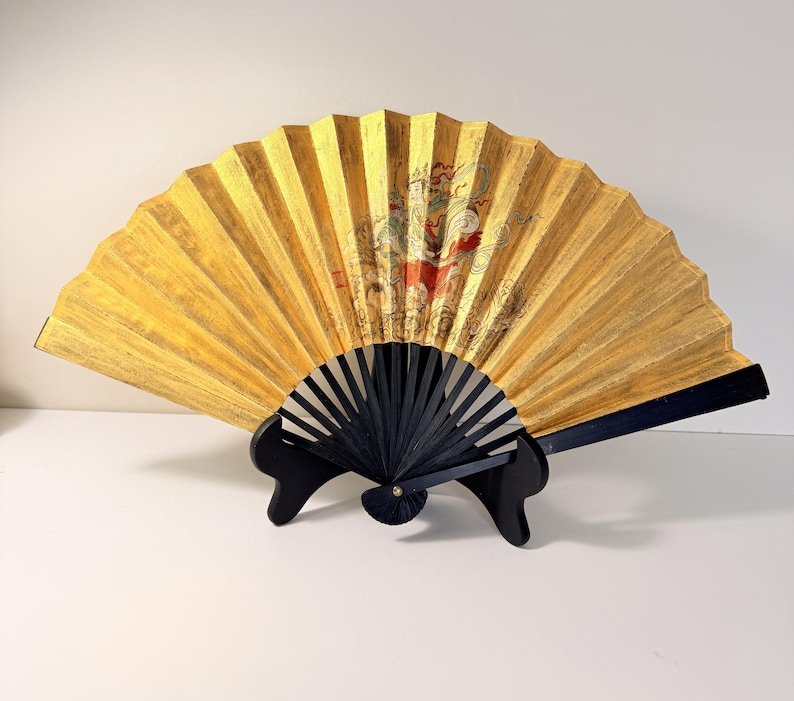 May include: A decorative folding fan with a gold-colored surface and a black handle. The fan features an ornate design with red and gold accents. The fan is displayed on a black stand.