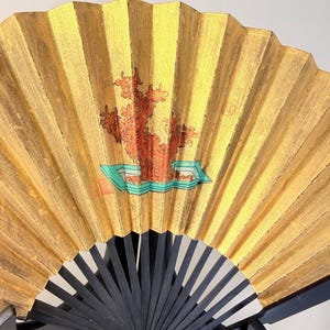 May include: A gold-colored, folding hand fan with a black handle. The fan features a painted design of a red tree-like shape on a turquoise base. The fan is made of pleated paper or fabric.