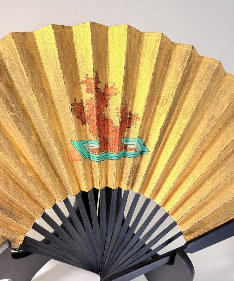 May include: A gold-colored, folding hand fan with a black handle. The fan features a painted design of a red tree-like shape on a turquoise base. The fan is made of pleated paper or fabric.