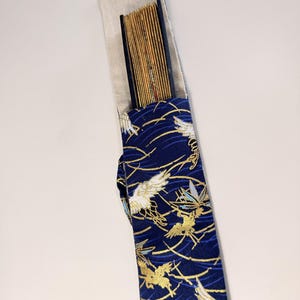 May include: A traditional Japanese fan in a navy blue case. The case features a pattern of gold and white birds and gold wave designs. The fan is made of thin, light-colored wooden slats.