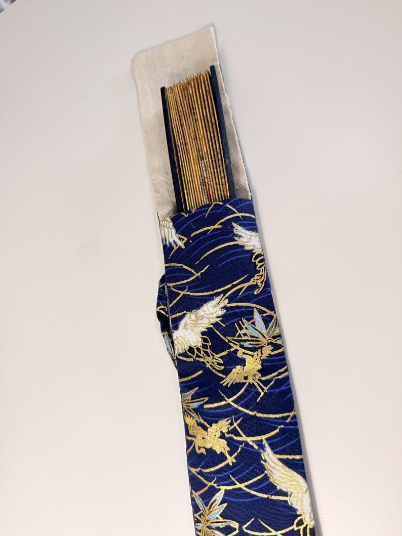 May include: A traditional Japanese fan in a navy blue case. The case features a pattern of gold and white birds and gold wave designs. The fan is made of thin, light-colored wooden slats.