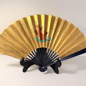 May include: A decorative gold-colored hand fan with a black handle and a black stand. The fan features a painted design with red and green accents. The fan is displayed open, showcasing its intricate design and craftsmanship.