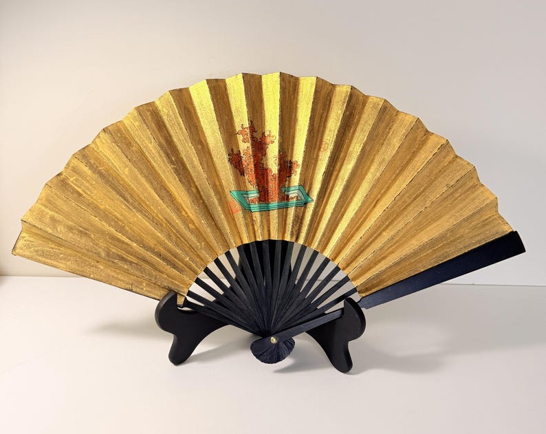 May include: A decorative gold-colored hand fan with a black handle and a black stand. The fan features a painted design with red and green accents. The fan is displayed open, showcasing its intricate design and craftsmanship.