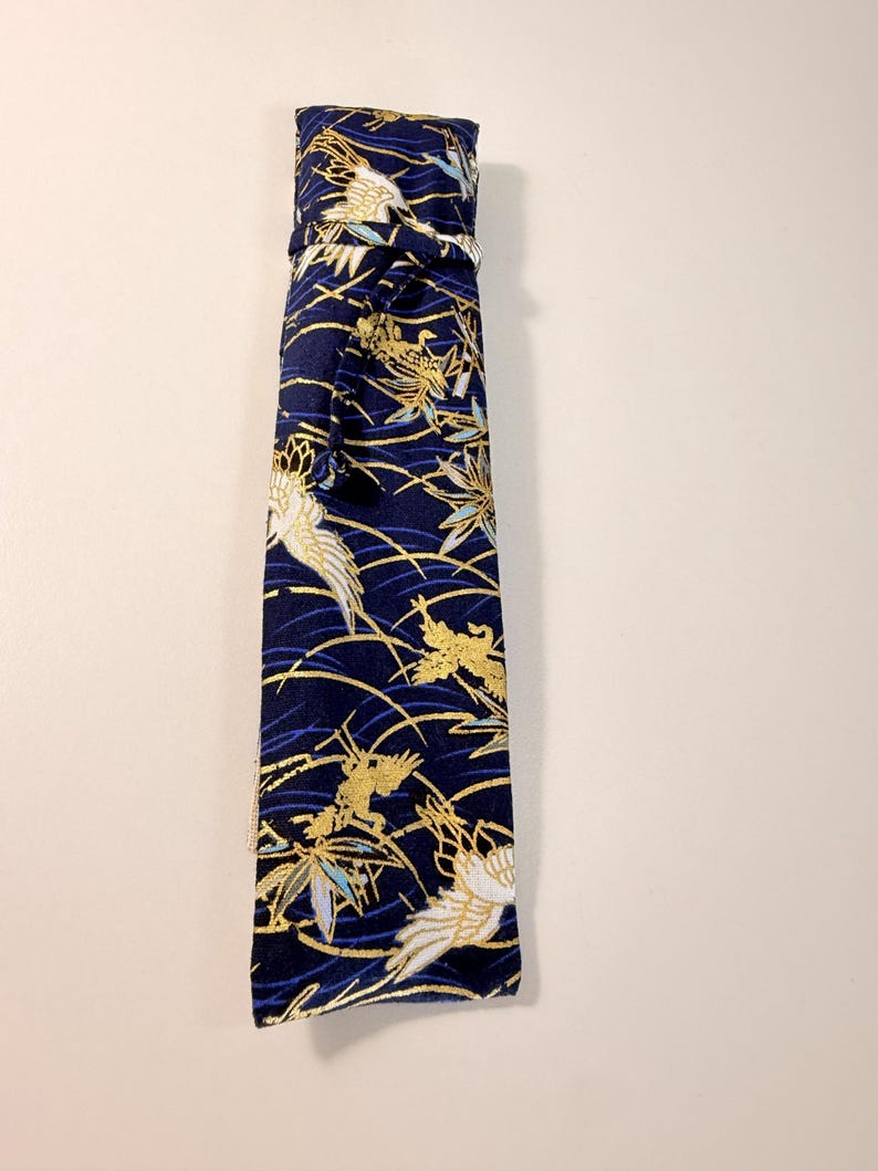 May include: A navy blue fabric item with a repeating pattern of gold and white birds and foliage. The item is tied at the top with a matching fabric tie. The design includes gold, white, and blue accents.