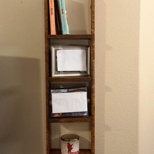 May include: A dark brown wooden ladder shelf with three levels. Two books sit on the top shelf, a framed picture on the middle, and a candle on the bottom. The shelf is mounted on a beige wall. The shelf's height is approximately 90 cm.
