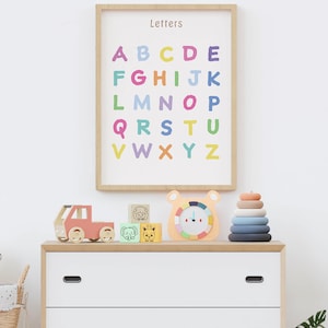 May include: A wooden framed print displaying the alphabet in various colours, with the word "Letters" at the top. A white chest of drawers holds a toy truck, animal blocks, a bear clock, and a ring stack. The room has a bright and cheerful atmosphere.