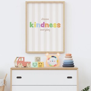 May include: A framed print with the words "choose kindness everyday" in colourful letters hangs above a white chest of drawers. The chest is topped with wooden toys, including a truck, animal blocks, a clock, and a stacking ring toy.