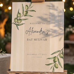 Op de afbeelding: Een houten ezel toont een welkomstbord voor Alexandra's Bat Mitzvah. Het bord heeft illustraties van olijftakken en de tekst "Welcome to Alexandra's Bat Mitzvah" en evenementdetails. De scène speelt zich buiten af met planten en lantaarns.
