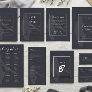 May include: A collection of dark gray event stationery, including invitations, a seating plan, and table cards. The designs feature white text and decorative floral elements. Ribbons and burlap accents complement the stationery.