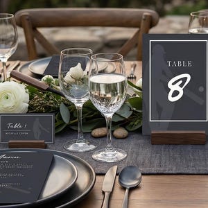 May include: A beautifully set table for an outdoor event. The table features dark gray plates, silverware, and wine glasses. A centerpiece of greenery and white flowers adorns the table. A table number card displays the number 8.