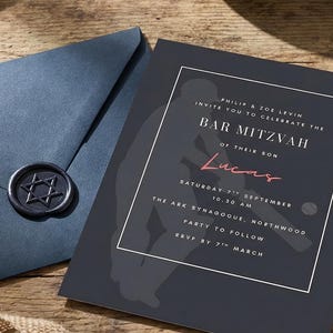 May include: A Bar Mitzvah invitation with a navy blue envelope and wax seal, a baseball, and a baseball glove are arranged on a wooden surface. The invitation reads "Bar Mitzvah Lucas". A navy blue kippah and prayer shawl are also present.
