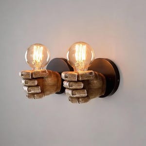 May include: Pair of wall sconces featuring a hand-shaped design holding a vintage-style light bulb. The hands are made of wood-like material, mounted on black circular backplates. The bulbs emit a warm, inviting glow.