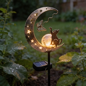 Könnte beinhalten: Ein solarbetriebener Gartenstecker mit einem Monddesign mit Sternaussparungen, einem Schmetterling und einer Katzen-Silhouette. Der Mond hält eine leuchtende Glaskugel. Der Stecker steht in einem Garten mit grünem Laub.