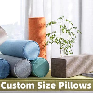May include: An assortment of custom-sized pillows in different shapes and colours. The image shows cylindrical and rectangular pillows in shades of blue, grey, and brown, with an orange cylindrical pillow in the background. The text "Custom Size Pillows" is at the bottom.