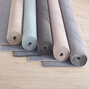 Puede incluir: Alfombras enrolladas en tonos beige, gris y azul, dispuestas sobre un suelo de madera clara. Se aprecia la textura y la variedad de colores de las alfombras. Los bordes de las alfombras son visibles, mostrando su construcci&oacute;n.